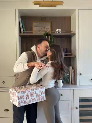 A couple embracing in a kitchen setting. The man is holding a decorated gift box, and both are smiling. The background includes white cabinets, shelves with books and decor, and a glimpse of a window with natural light.