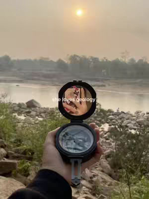 The cover image captures a scenic outdoor shot with a hand holding an open compass, reflecting a partial view and a tattoo. The background reveals a body of water, hills, and a bright sun in a hazy sky. Text overlay reads 'she loves geology'.