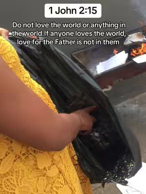 The image shows part of a person, arm and torso, wearing a yellow lacy dress, standing in front of what appears to be a large black trash bag, the hand is pointing inside to something. In the background, there is some fire in a metallic container. There is some text overlaid on top.