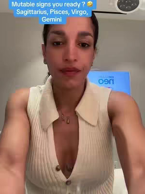 A woman with brown skin and dark hair gazes directly at the camera, offering a slight smile. She wears a cream-colored collared top. The background is a blurred interior, with some lighting and a screen visible.