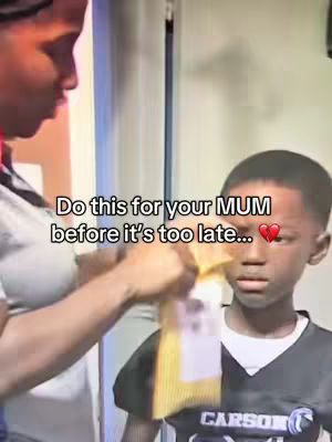 A candid shot shows an adult hand holding a small, yellow item as they stand in front of a young boy, possibly their son, who has a somewhat apprehensive look. The text overlay states: "Do this for your MUM before it's too late...", with a heart symbol.