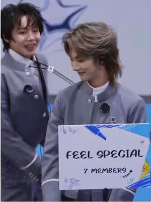 Two young men are standing side-by-side, appearing to be in a studio or on a stage. One person is smiling, while the other is looking down, holding a sign that reads "FEEL SPECIAL" with additional text. They are wearing grey jackets and white shirts.