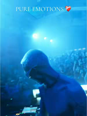 The image captures a blue-toned scene of a concert or music event. A figure, possibly a performer, is seen from behind wearing headgear and sunglasses, looking towards a blurred, illuminated crowd. Text overlays at the top read 'PURE EMOTIONS' followed by a heart emoji.