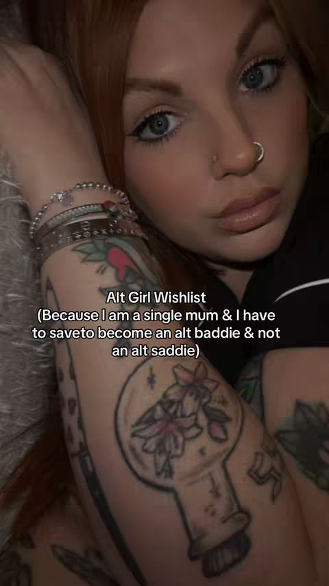 A close-up selfie featuring a person with red hair lying down, their arm with multiple tattoos in the foreground. Text overlays the image, and the person has a nose ring and stylish eye makeup.