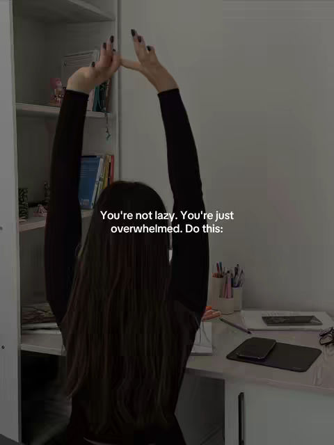 A person with long, dark hair is seen from the back, sitting at a desk with their arms raised above their head. The person is wearing a black long-sleeved shirt. In the background, a wall, a shelf, and a desk are visible