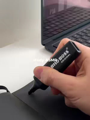 A hand holding a black marker, writing on a black notebook, with a laptop in the background. The laptop screen displays an image. The scene is on a white surface.