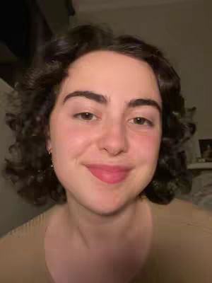 A close-up shot of a young woman with dark curly hair. She is smiling subtly, her pink lips slightly parted. Her skin appears fair, with some natural redness. The background is blurred and indistinct, but seems to be inside a building. She is wearing a neutral-toned shirt. The lighting is soft and looks natural.