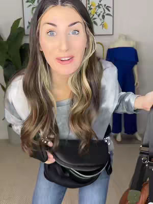 A woman with long brown hair is centered, looking at the camera. She's holding a black bag. The background includes a plant, some framed art, and a blue jumpsuit on a mannequin.