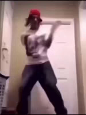 A person wearing a red cap and white shirt, dancing in a room. The room has white walls and a white door. The person is silhouetted and the image is low-resolution.