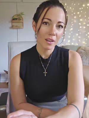 A woman with brown hair and brown eyes is looking directly at the camera with a slight smile. She is wearing a black sleeveless top and a silver necklace with a cross pendant. The image is taken against a softly lit background. The overall composition is balanced, with the woman as the central focus and the background providing a subtle backdrop.