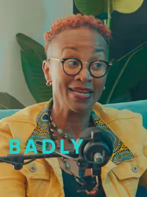 A woman wearing glasses and a yellow jacket is positioned in front of a microphone. The word "BADLY" is overlaid across the center of the image in a turquoise font.
