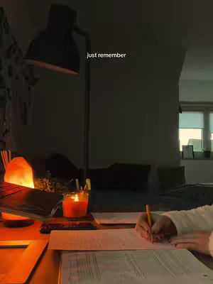 A dimly lit workspace at night. Shows a person's hand writing on paper with a pencil, a lit candle, a lamp, a laptop, and a salt lamp.