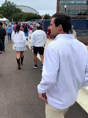 The cover image shows a man walking on a paved pathway, appearing to be at an outdoor event or gathering. He's facing away from the camera, looking towards a crowd of people in the distance. The man is wearing a white top and khaki pants. In the background, other people are visible, and a large building is partially visible. The composition gives a sense of movement and activity. The colors are natural, with an emphasis on the white of the man's top and the background crowd's attire.