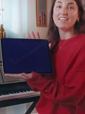 A woman wearing red holding a tablet with a blank screen, smiling at the camera. In the background is a piano and a framed picture.