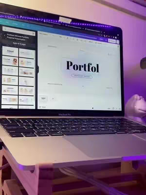 A laptop displaying a Canva interface with a presentation titled 'Portfol' on its screen. The laptop is supported by a wooden stand. The overall lighting is a mix of natural and purple ambient light.