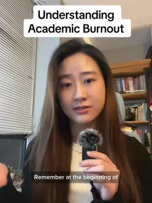 A woman with long brown hair is holding a microphone and speaking. A text box in white is at the top, and more text appears in a black box at the bottom. The background has shelves with books.