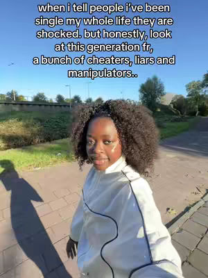 A young Black woman with curly hair is standing outdoors on a sunny day. She is wearing a white jacket with black trim and looking towards the camera with a slight smile. The background shows a sidewalk, a street, some trees, and a blue sky. Text is overlayed on the image.