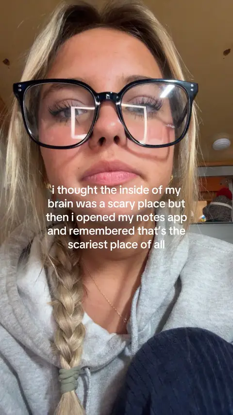 A close-up shot of a young woman wearing glasses, her face centered in the frame. She has blonde hair styled in a braid on the side and is wearing a grey hooded sweatshirt. The background is a warm, neutral tone.