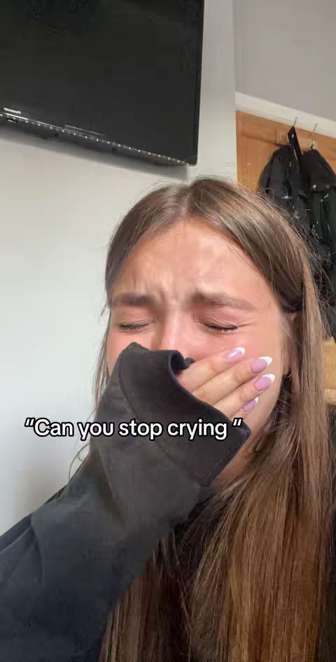 A close-up shot of a young woman crying, her face partially obscured by her hand. The background is a blurred interior with a television and a door visible.