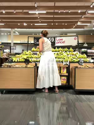 A woman in a white skirt and tank top is standing in a grocery store, facing away from the camera. Behind her are displays of bananas and store signage.