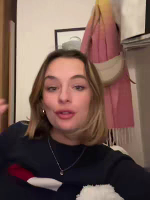 A young woman with shoulder-length brown hair, wearing a dark navy blue sweatshirt with a red, white, and blue emblem, talks directly to the camera. The background is a room with a light-colored wall and a picture frame. A pink scarf and a cream-colored purse hang on a clothing rack behind her. The lighting appears natural, and the overall composition is a medium shot focusing on the woman's face and upper body.