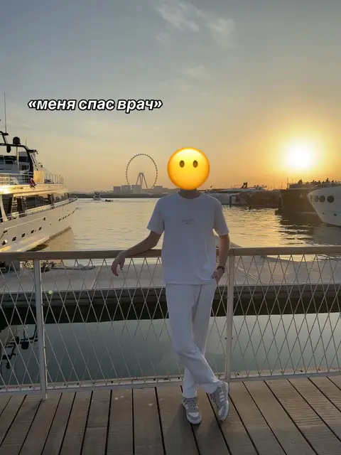 A person with a yellow smiley face emoji covering their head stands on a wooden surface. They are wearing a white t-shirt, white pants, and gray sneakers. The background shows a body of water, boats, and a Ferris wheel. The sky is a gradient of orange and blue, suggesting a sunset. Text overlayed in a bubble reads, “I was saved by the doctor” in Cyrillic script.