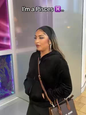 A woman is standing in a hallway with a large mirrored or reflective surface to her left. She is wearing a black hoodie and a black headband. She has a shoulder bag and a purse. The text overlay reads 'I'm a Pisces ♓️..'