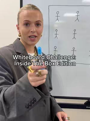 A woman with blonde hair stands in front of a whiteboard, holding a blue marker and looking at the camera. Behind her is a whiteboard with stick figures drawn on it, within a boxed outline. The woman is wearing a gray blazer, gold hoop earrings, and rings. The background is a white wall and the overall lighting is bright.