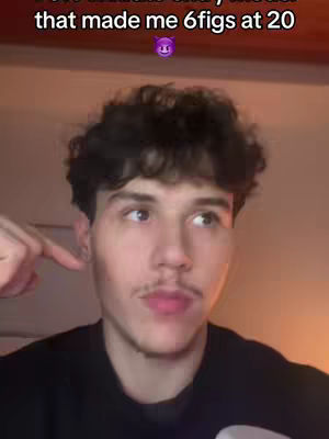 A young man, wearing a black shirt, is looking slightly off-camera with a finger pointed toward his ear. Text overlay at the top says, "that made me 6figs at 20" with a purple devil emoji. The background is a blurred interior.