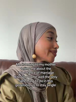 A woman wearing a gray hijab and an olive green jacket is seen in a close-up shot. The image is warm-toned, with the woman's skin appearing smooth. Text overlay appears. The background is a light-colored wall.