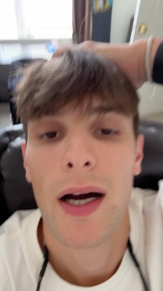 A young man with brown hair and a white shirt is seen in a close-up shot, looking directly at the camera with an open mouth. The background is blurred but appears to be a domestic setting. His hand is touching his hair.