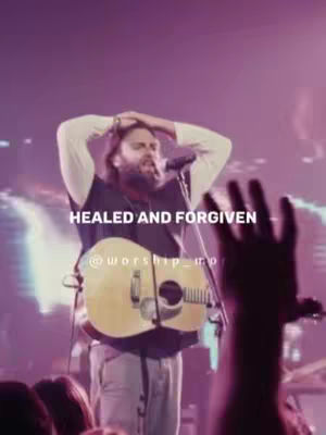 A man with a guitar and microphone is on stage, bathed in purple and white stage lighting. Text overlays read 'HEALED AND FORGIVEN' with social media handles.