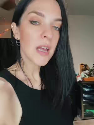 A woman with dark hair and fair skin is shown in a medium shot. She is looking towards the camera with her mouth open as if speaking. The lighting appears to be natural. She is wearing a black top and small earrings.