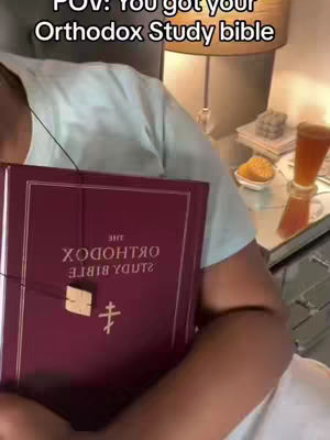The video features a person holding a large burgundy-colored book with a gold emblem and a cross symbol. The person appears to be inside, with a blurry lampshade, a mirror, and a glass of liquid visible in the background. The text overlay says: POV: You got your Orthodox Study bible