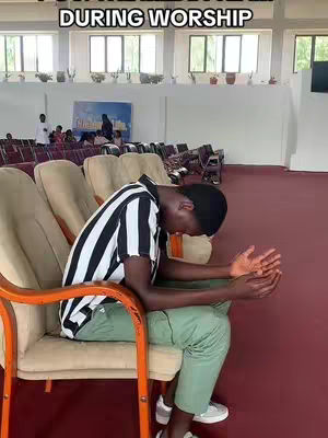 A young Black man wearing a black and white striped collared shirt and olive green pants sits in a cream-colored chair in a church. He has his head bowed, and his hands are clasped together in front of him, as if in prayer or deep thought. The background includes more seating, a few people, and windows letting in soft light.