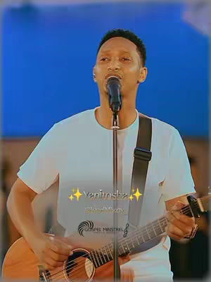 A man is shown playing a guitar and singing into a microphone. The background is a solid blue, and text overlays the image, which includes the title of the song. The man is wearing a white t-shirt, and has a guitar strap over his shoulder. The lighting appears to be natural, and the overall composition is centered.