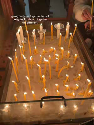The image shows a close-up view of many lit candles set in a sandy enclosure. The candles vary in height and are positioned closely together, with the flames flickering brightly. In the background, a few additional, taller candles are visible, and a pair of hands holding a candle can be seen on the right side, suggesting an active setting like a church.