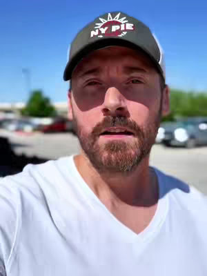 The cover image features a close-up shot of a man wearing a baseball cap with a logo. He appears to be outdoors on a sunny day, with a blurred background of a parking lot and some greenery. The man is looking directly at the camera with a slightly open mouth, suggesting he is speaking.