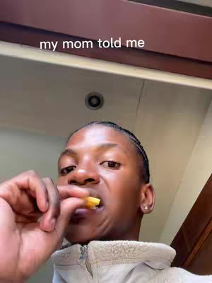 A person with a dark complexion is shown from the chest up, biting into a french fry. The individual is eating fries, with a slight wrinkle in their nose. Their hair is styled back in small braids. The person wears a beige, zip-up jacket. Behind them is a beige ceiling and a visible light fixture.