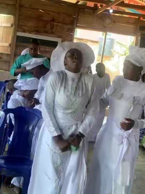 The image shows a group of people in a church setting. The main subject, a woman, is in the center, wearing a white dress and hat, with arms crossed. Other people in white clothing are in the background. The composition suggests a religious gathering or ceremony.