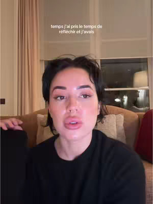A woman with short dark hair and makeup is centered in the frame, with a light-colored background. She appears to be speaking and is wearing a black shirt. Text overlay in the image. the focus is on her face.