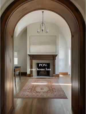 An interior shot framed by a dark wooden archway. The view leads into a bright, open room with hardwood floors. A patterned rug sits centered on the floor in front of a fireplace and a large piece of art that dominates the wall. The lighting is soft, and the space feels clean and inviting.