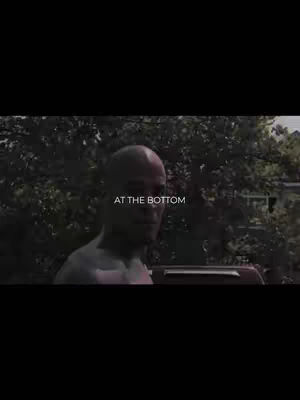 The image is a medium shot of a man, likely outdoors. The man has a shaved head, and the shot appears to be taken from around the chest up, with a dark background of dense trees and foliage. The text overlay, 'AT THE BOTTOM,' is positioned centrally below the man's face.