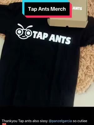 A black t-shirt with the Tap Ants logo is displayed, along with a box. The shirt is centered, and the logo is a white illustration of an ant with glasses and the words "TAP ANTS" in large, white letters. A cardboard box is partially visible in the background, and the overall setting has a warm, fuzzy texture.