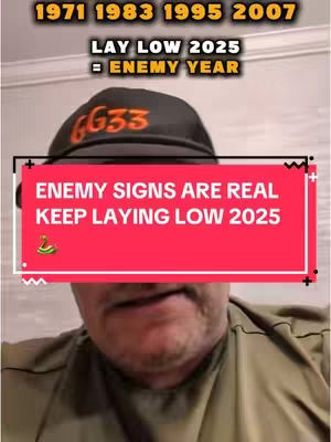 The image is a selfie-style shot featuring a man with a partially visible face. The man is wearing a black cap with the text 'GG33' in orange. A red rectangular overlay covers the lower part of the cap and the upper part of the man's face, displaying the text 'ENEMY SIGNS ARE REAL KEEP LAYING LOW 2025'.  Above the overlay, in a yellow font, appear the numbers 1971, 1983, 1995, and 2007, with the text 'LAY LOW 2025 = ENEMY YEAR'. He is wearing a patterned green shirt. The background is plain.