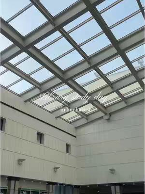 An upward shot of a building's interior roof displaying glass panels and metal beams, creating a grid-like pattern against a light blue sky. The lower portion of the image shows a white wall with evenly spaced rectangular windows and what appear to be lighting fixtures. Sunlight shines through.