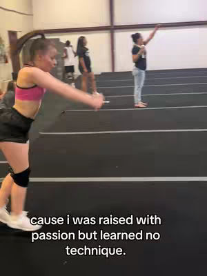 A young woman in athletic attire is mid-flip in a large gymnasium. She is wearing a pink sports top and black shorts, with a knee brace on one leg. Other people in the background are watching. The floor is black with white lines and the walls are white.