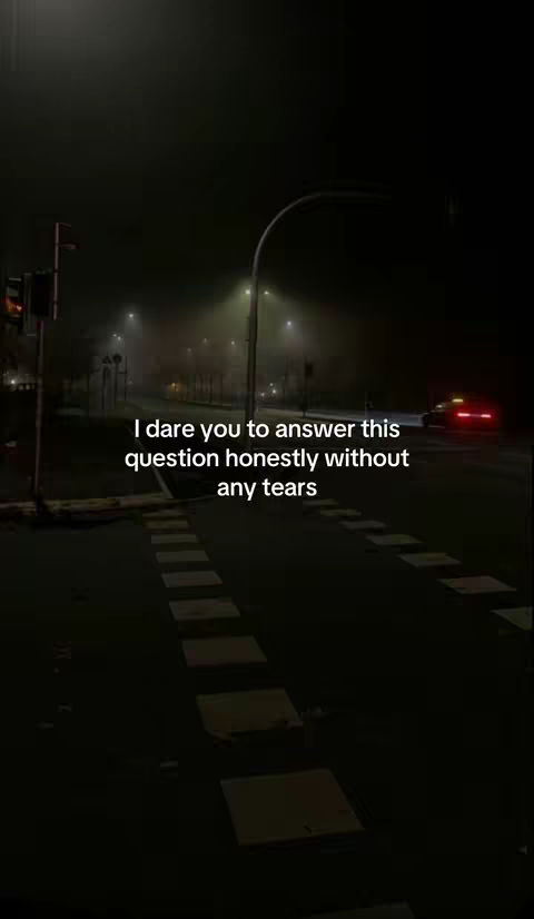A nighttime scene on a city street. The foreground shows a pedestrian crossing with white stripes. The street is dark, with streetlights casting a yellowish glow and fog. In the background, a red car is visible. The sky is obscured by fog, adding to the mood.