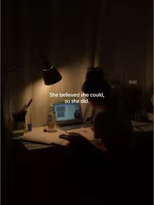 The image is a dimly lit study scene. A person is seated at a desk with a laptop, papers, and a lamp casting light. Text overlayed reads: 'She believed she could, so she did.'