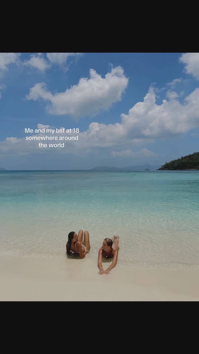 The cover image shows two young women lying on their stomachs in shallow, clear water at the edge of a white-sand beach. The sky above is a brilliant blue dotted with fluffy white clouds. There is a lush green island and a sailboat in the distance. Text overlayed on the image reads, "Me and my bsf at 18 somewhere around the world". 